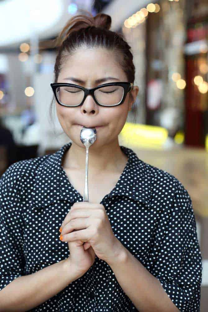 Woman in black shirt and white polka dots and black glasses with spoon in mouth tasting for flavor. | MakeSauerkraut.com