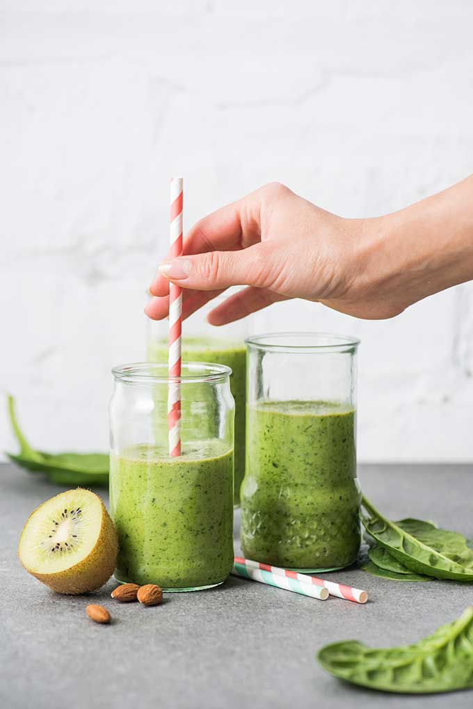 Glasses of green smoothie with two striped red and white straws, and one blue and white straw and half a kiwi and some nuts to the side as decorations. | MakeSauerkraut.com