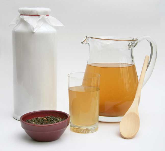 Jug, glass and mental container containing kombucha, bowl with dried tea herbs and a wooden ladle. | MakeSauerkraut.com