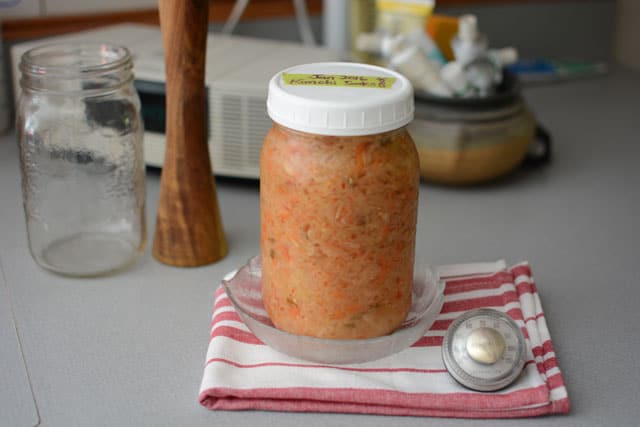 Jar of sauerkraut in a small bowl over striped red and white cloth and a round thermometer at the side, an empty jar and wooden tamper in the background. | MakeSauerkraut.com