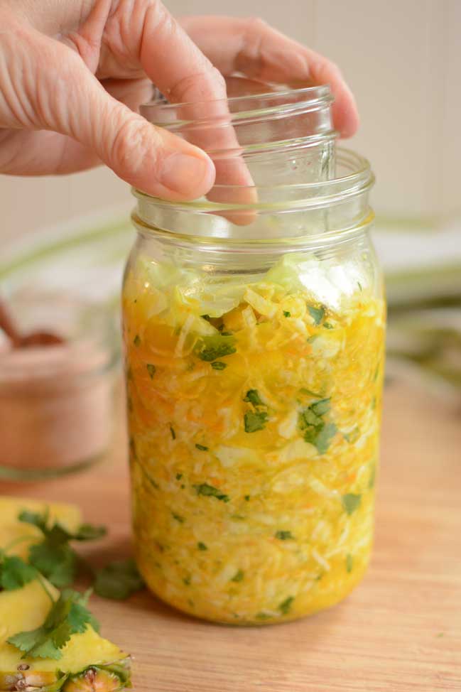 A hand inserting a small jelly jar into a glass jar of sauerkraut as a fermentation weight. | MakeSauerkraut.com