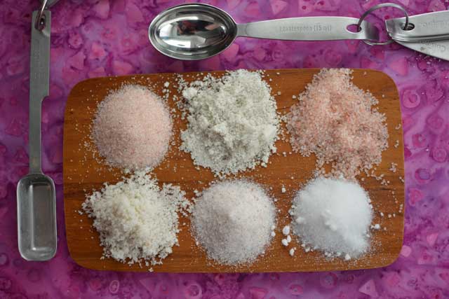 Six different kinds of salt in small piles on top of a wooden chopper board and a metal scoop the to the left and measuring spoons on top. | MakeSauerkraut.com