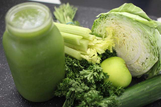 An opened jar of celery juice and fresh celeries, apple, cucumber and half a cabbage. | MakeSauerkraut.com