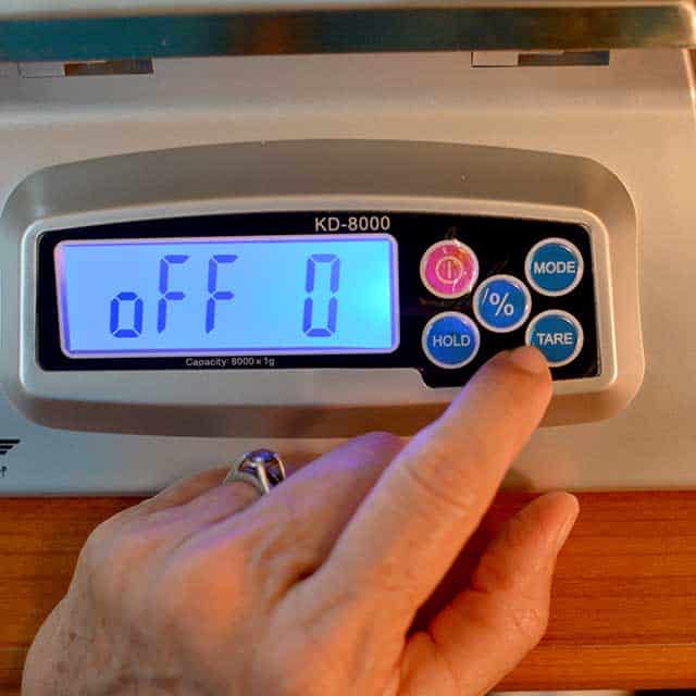 Fingers reaching to press "tare" on the MyWeigh KD-8000 digital scale with monitor showing "off 0". | MakeSauerkraut.com