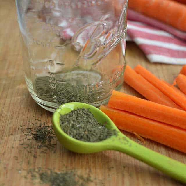 Green spoon with dried dill, a jar with some dried dill inside and some carrot sticks to the side. | MakeSauerkraut.com