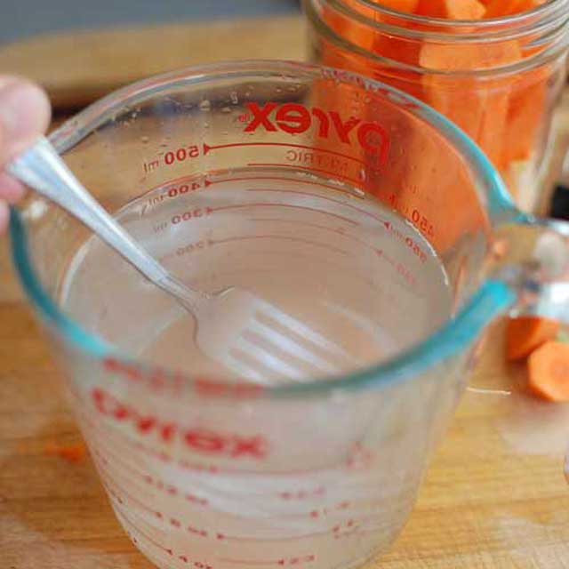 A large measuring bowl with clear brine and a fork mixing it and a jar filled with carrot sticks in the background. | MakeSauerkraut.com
