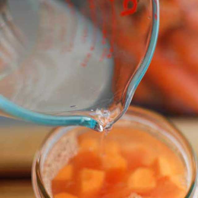 Measuring cup pouring brine over jar of carrot sticks. | MakeSauerkraut.com