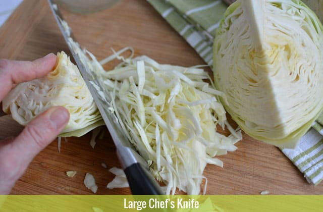 Hand holding a small portion of a cabbage with a large chef's knife doing the slices and another quarter of a cabbage. | MakeSauerkraut.com