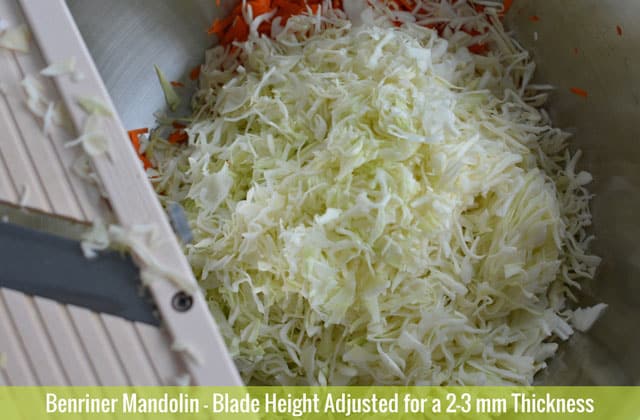 A pile of thinly cut cabbages in a metal container and a Benriner Mandolin at the left side of the image with clumps of cabbages. | MakeSauerkraut.com