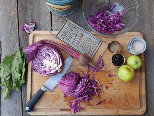 Ingredients for Ruby Red Sauerkraut with half a head of red cabbage near a knife with sliced cabbages at the side, metal grate, two green apples, small bowl of dried cranberries, herbs, jar of salt placed over wooden cutting board and big glass bowl with sliced red cabbages. | MakeSauerkraut.com