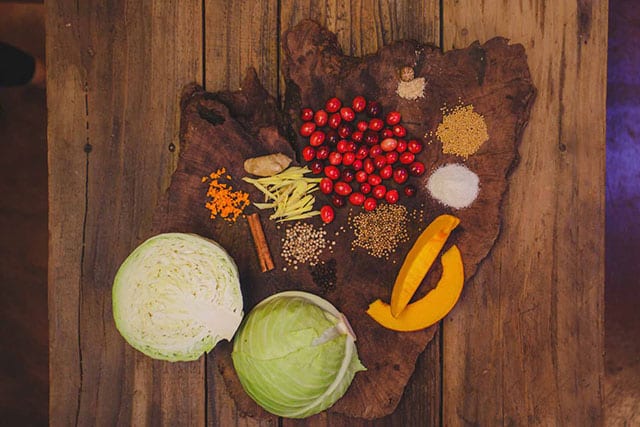 Two half heads of cabbages, two slices of pumpkin and other ingredients for Pumpkin Cranberry Sauerkraut over wooden slab. | MakeSauerkraut.com