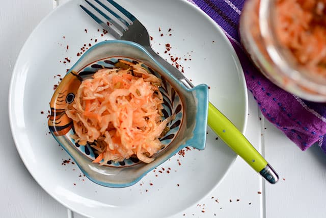 Top view of a serving of kimchi-styled sauerkraut in a fish shaped bowl over white plate with fork with green handle at the top right. | MakeSauerkraut.com