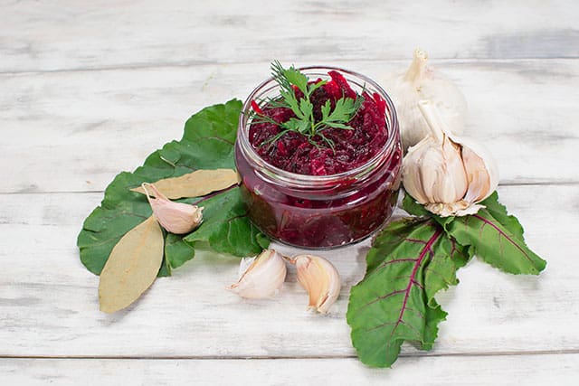 Beet relish in a small glass container surrounded by cloves of garlic and big beet leaves and dried bay leaves. | MakeSauerkraut.com