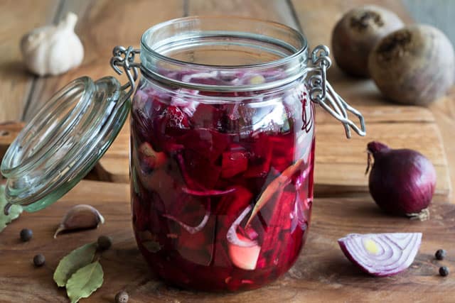 A glass jar with the metal clamp lock with diced beets in brine with onions and some cloves of garlic scattered at the background. | MakeSauerkraut.com