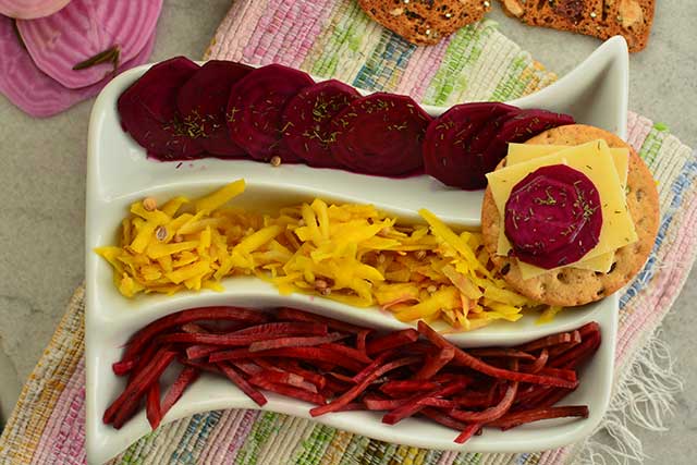 A three sectioned spiral white plate with a biscuit on its top right corner and fermented beets and cheese. The top section of the plate has a fermented beets cut circularly, the middle part has some yellow vegetables, and the bottom section has some fermented bets sliced like French fries. | MakeSauerkraut.com