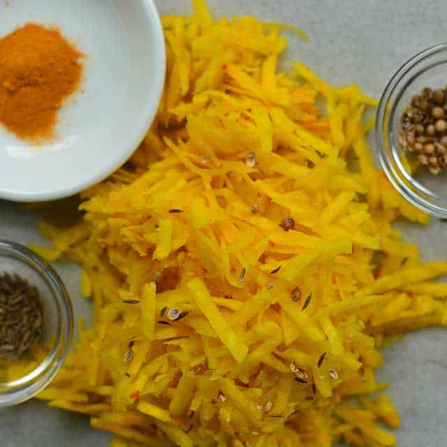 A pile of grated yellow beets with a small white plate with turmeric powder and two small glass bowls on the bottom left and top right of the picture with herbs. | MakeSauerkraut.com