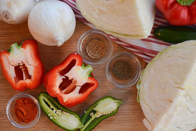 Top view of ingredients for Baseball Park Sauerkraut Recipe. like two halves of red bell pepper, two halves of green peeper, two heads of garlic, three small bowls with herbs and spices, and quarters of cabbages.  | MakeSauerkraut.com