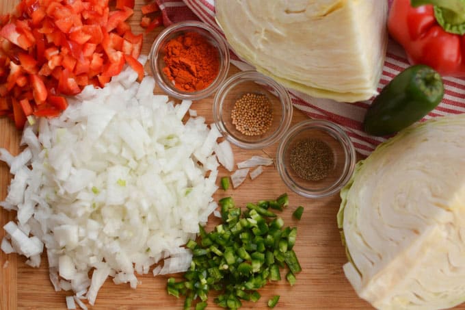 Top view of ingredients for Baseball Park Sauerkraut Recipe with chopped onions, green pepper and red bell pepper, three small bowls with herbs and spices, and quarters of cabbages. | MakeSauerkraut.com