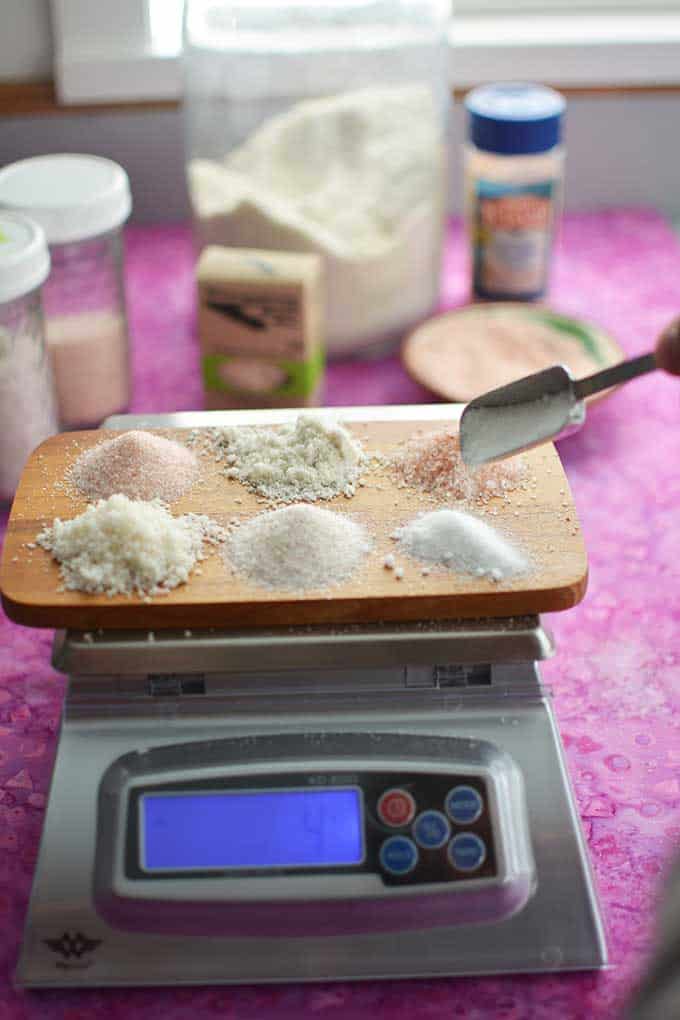 Different types of salt in small batches in wooden board being weighed over a digital scale. | MakeSauerkraut.com