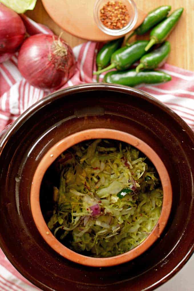 Top view of sauerkraut inside brown, water sealed fermentation crock with red and white cloth, red onions, green peppers, and a small bowl of herbs in the background. | MakeSauerkraut.com