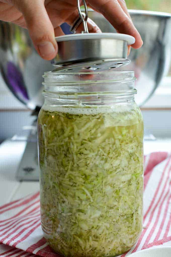 Fingers inserting the fermentation lid by Kraut Source into a jar of sauerkraut with the Pickle Plate in clear view at the bottom of the lid. | MakeSauerkraut.com