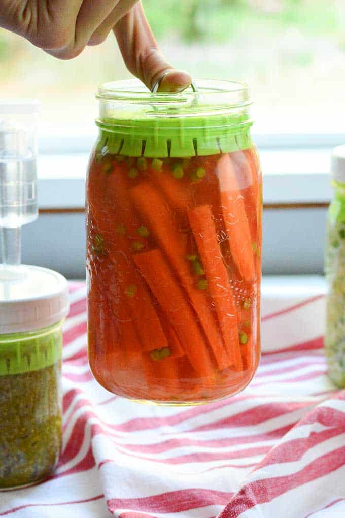 Finger lifting a jar of fermented carrots using the a small hook attached to the Pickle Pusher fermentation weight. | MakeSauerkraut.com