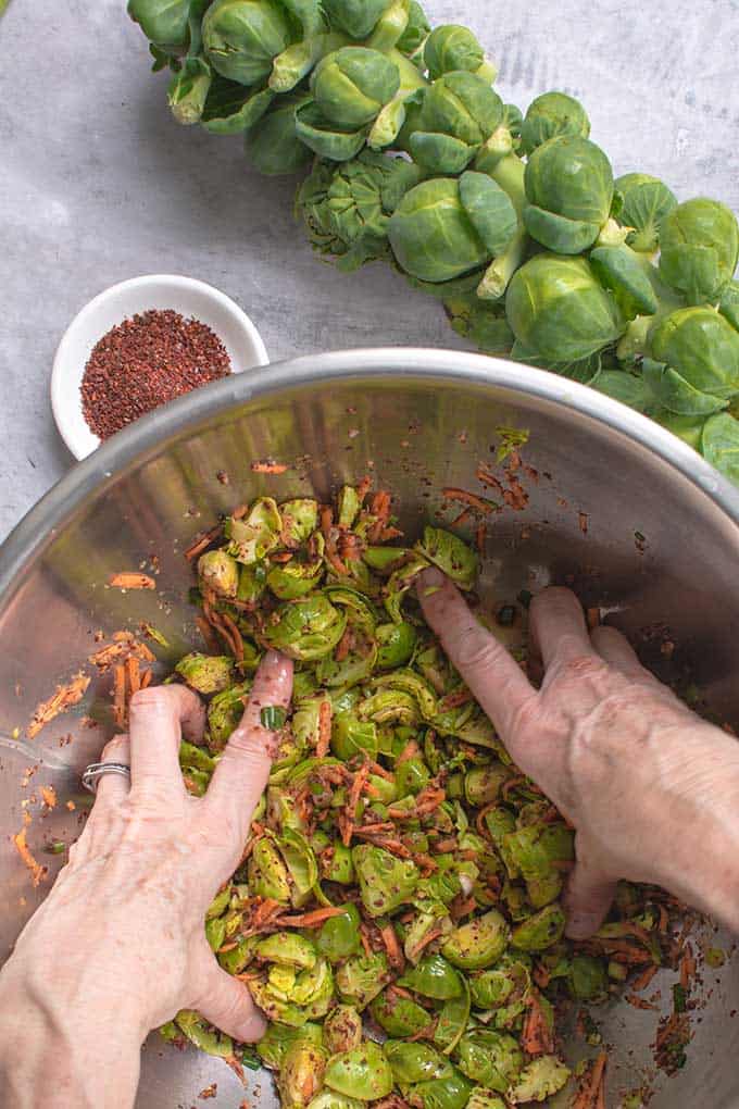 Hands mixing a brussels sprouts in a metal bowl with fish sauce and Gochugaru, a fresh stalk at the upper right corner of the bowl and a saucer of Gochugaru to the upper left. | MakeSauerkraut.com