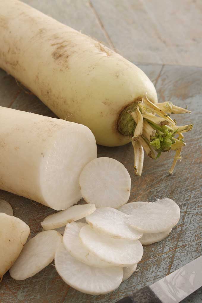 Two fresh daikon radishes, one showing the top head with leaves and the other being sliced. | MakeSauerkraut.com