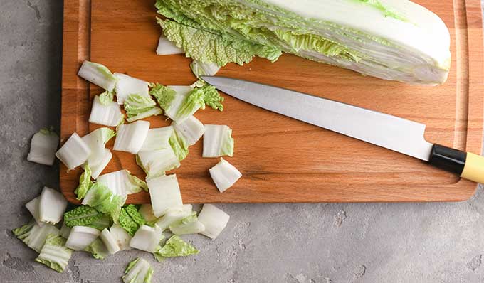 Half of the napa cabbage with a knife and small square cuts of cabbage on top of wooden cutting board. | MakeSauerkraut.com
