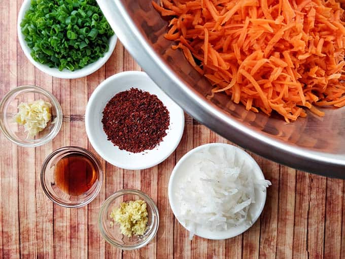 Carrot Kimchi ingredients prepped and ready for mixing with piles of grated carrots inside a big metal bowl and herbs and spices in small and white bowls. | MakeSauerkraut.com