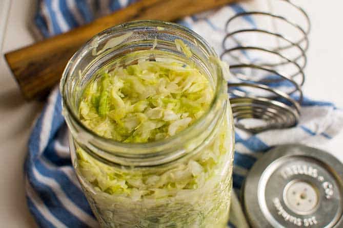 Opened jar of sauerkraut with the PickleHelix and metal lid behind it over striped blue and white cloth. | MakeSauerkraut.com