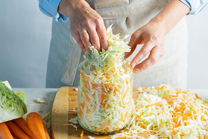 Woman packing a jar with sliced cabbage and grated carrot mixture for making sauerkraut.