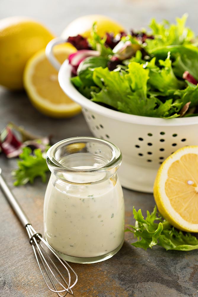 Image with the central focus being a small glass jar with of ranch-style dressing, a small whisk, and half a lemon slice, and white strainer bowl with vegetable leaves. | MakeSauerkraut.com