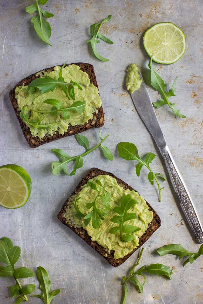 Two avocado toast on a metal counter with a butter knife at the side with lime and aragula leaves scattered all over. | MakeSauerkraut.com
