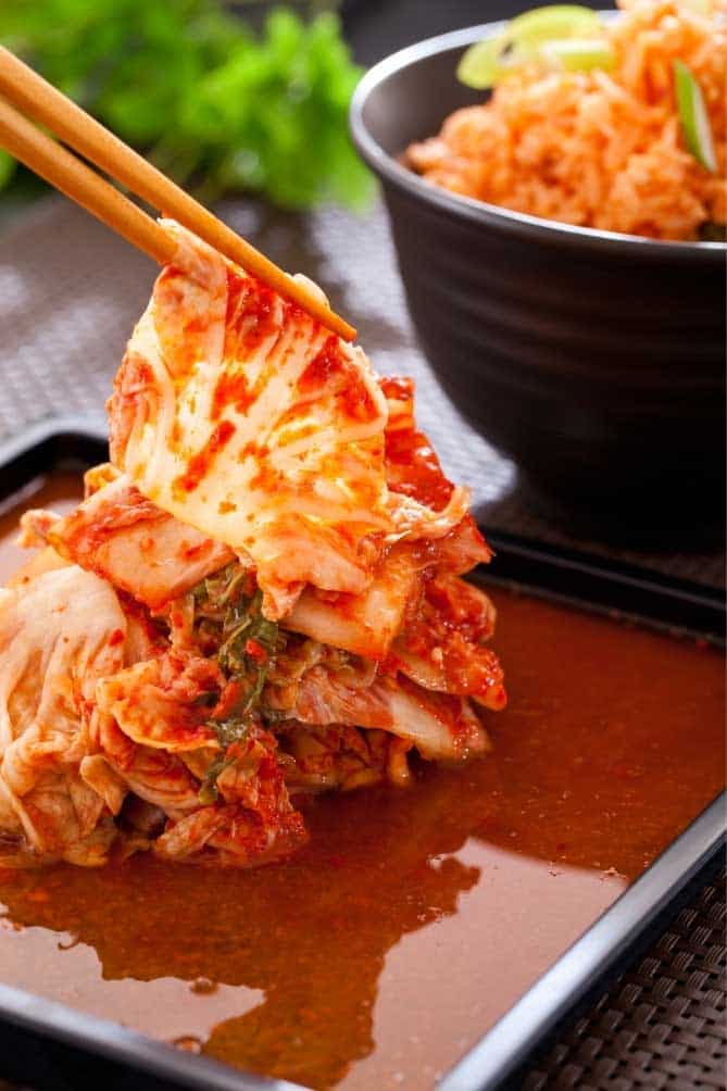 Serving of napa cabbage kimchi being picked up by wooden chopsticks and a black bowl with more servings. | MakeSauerkraut.com