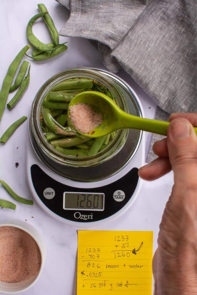 A green spoon with salt added to a jar of beans on a scale. | MakeSauerkraut.com