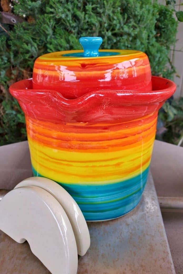 fermentation crock A rainbow-colored ceramic fermentation crock with a red lid and blue knob, accompanied by two white ceramic weights. | makesauerkraut.com